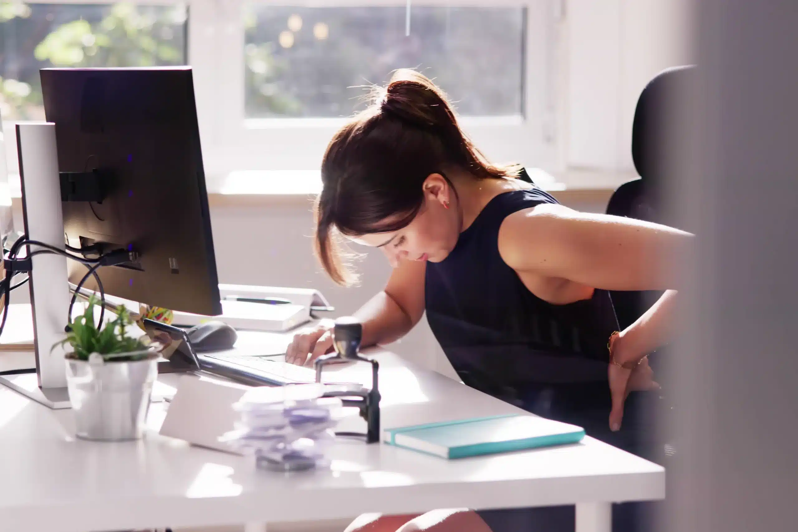 Woman sitting in office with bad posture and back pain, showing effects of prolonged sitting on spinal health.